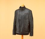 Black leather jacket displayed on a stand against a beige background. The jacket features multiple zippers and a high collar, conveying a sleek, stylish look.