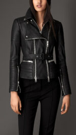 A woman wears a stylish black leather biker jacket with quilted sleeves and silver zippers, paired with black pants. The tone is edgy and chic.