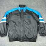 The NFL Black Blue Carolina Panthers Bomber Leather Jacket features blue and white shoulder stripes, a blue embroidered Panthers logo on the left chest, and is shown laid flat on a grey surface. Ideal for any NFL leather jacket collection.