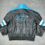 The NFL Black Blue Carolina Panthers Bomber Leather Jacket features black leather with blue shoulders, white sleeve stripes, and a large team logo on the back, displayed on a gray surface.