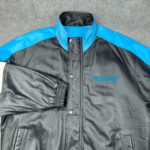 A NFL Black Blue Carolina Panthers Bomber Leather Jacket with blue shoulders and collar, snap buttons, and Fubu Sports embroidered in blue on the left chest, is laid flat on a gray surface.