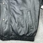 Close-up of the NFL Black Blue Carolina Panthers Bomber Leather Jacket with snap buttons, a leather-trimmed side pocket, and elastic hem, displayed on a black textured surface.